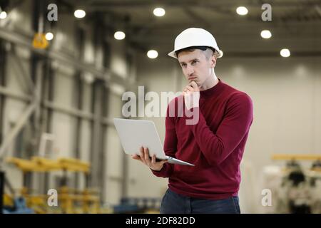 Ritratto di un manager di fabbrica maschile in un cappello bianco e un maglione rosso che tiene un computer portatile e un telefono cellulare. Controllo del processo di lavoro nel produttore di elicotteri. . Foto di alta qualità Foto Stock