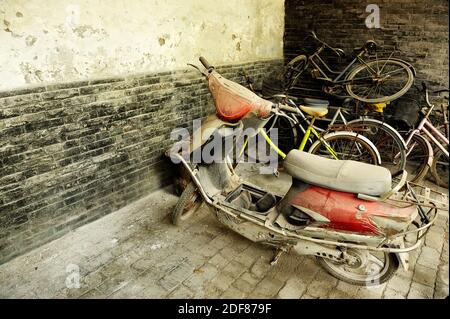 Vecchio rotto e abbandonato scooter coperti da polvere parcheggiato contro la parete a spiovente. Rusty pila di biciclette in background. Foto Stock