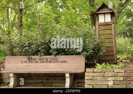 Una panca di parco di legno incisa con una citazione ispiratrice lungo una parete di ritegno di pietra, arbusti, alberi e una casa di luce di legno a Janesville, Wisconsin, Stati Uniti Foto Stock
