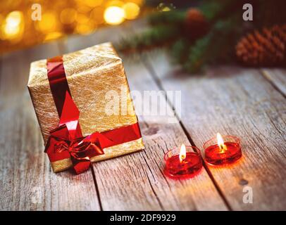 Scatola regalo d'oro e ramo di pino con coni con candele su tavolo di legno. Sfondo bokeh luci gialle. Vacanze di Natale e Capodanno a tema decoratio Foto Stock