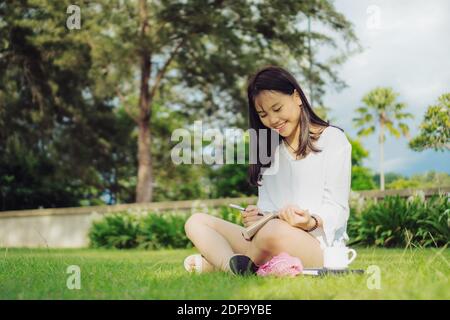 Sorridente giovane donna asiatica seduta con le gambe si incrociano su campo d'erba in un parco pubblico mentre scrive su diario. Fotografia stile di vita all'aperto. Foto Stock