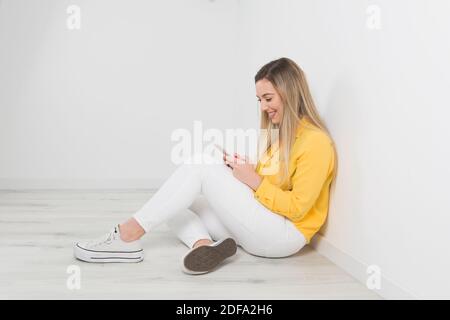 Una bella giovane bionda che sorride al suo smartphone seduto sul pavimento Foto Stock