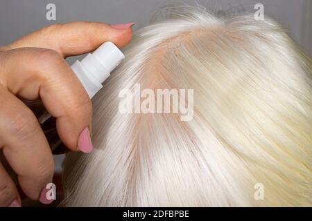 La donna mette sui capelli un tonico di idrolita da una bottiglia. Cura dei capelli a casa, pittura bionda, nutrizione e idratazione del cuoio capelluto Foto Stock