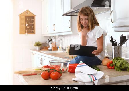 Ragazza piccola sulla panca della cucina piena di verdure che consultano ricette su un tablet Foto Stock