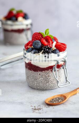 Due vasetti di muratori con budino di chia e lamponi freschi, fragole, mirtilli e menta su sfondo bianco con un cucchiaio di legno con semi di chia i Foto Stock