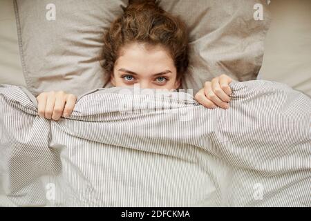 Vista ad alto angolo di una giovane donna che guarda la fotocamera nascondersi sotto la coperta nel letto Foto Stock