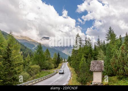 Un'auto si muove lungo una strada di montagna con un santuario votivo sul lato sotto un cielo drammatico, Solda, Alto Adige, Italia Foto Stock