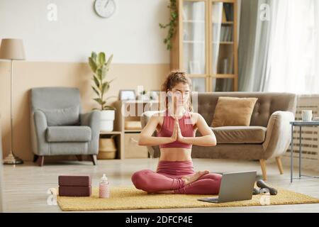 Giovane donna seduta sul pavimento e facendo yoga online utilizzando il suo computer portatile nella stanza di casa Foto Stock