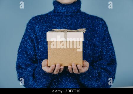 Buono di Natale, anno di nascita, Natale, banner di compleanno. Mani femminili in pullover blu che tiene scatola regalo artigianale con nastro su sfondo blu. Foto Stock
