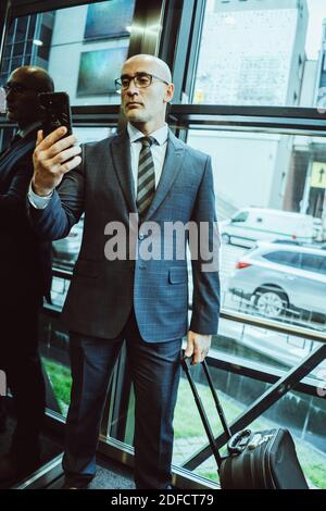 Uomo d'affari sicuro in tuta legge le informazioni su in smartphone. L'uomo tiene il telefono cellulare davanti a lui mentre si trova accanto alla finestra o all'ascensore Foto Stock
