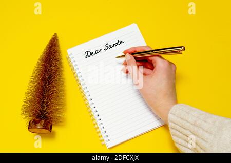 La mano femminile scrive la lettera a Santa su sfondo giallo. Albero d'oro di Natale e cara Santa scrittura a mano. Concetto di Natale. Foto Stock