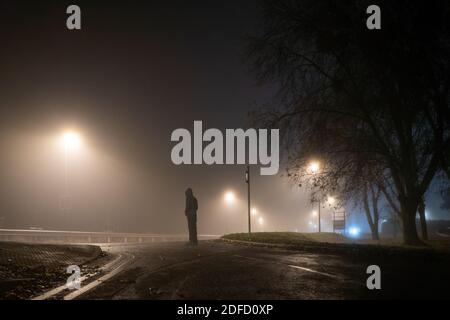 Guardando su una figura incappucciata in piedi su una strada con luci di strada su una suggestiva notte di inverni nebbioso. REGNO UNITO. Foto Stock