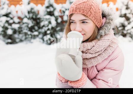 Ritratto di una bella giovane ragazza in bianco mittens lavorato a maglia, all'aperto, bere una bevanda calda, luminoso inverno day.woman sorridente e felice, macro, albero Foto Stock