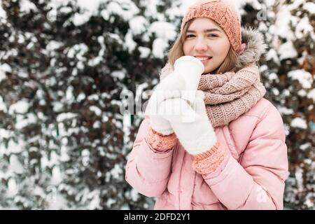 Ritratto di una bella giovane ragazza in bianco mittens lavorato a maglia, all'aperto, bere una bevanda calda, luminoso inverno day.woman sorridente e felice, macro, albero Foto Stock