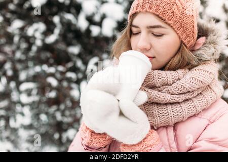 Ritratto di una bella giovane ragazza in bianco mittens lavorato a maglia, all'aperto, bere una bevanda calda, luminoso inverno day.woman sorridente e felice, macro, albero Foto Stock