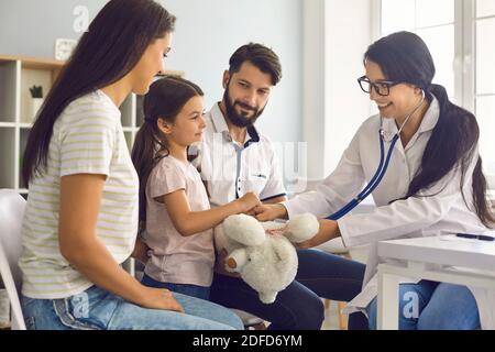 Il medico di famiglia ascolta con uno stetoscopio il bambino con i genitori seduti al tavolo della clinica. Foto Stock