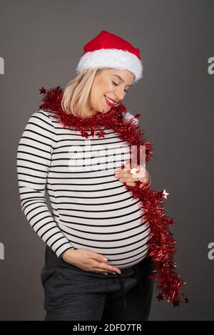 Bella donna incinta nel terzo trimestre indossa il cappello di Santa e rosso natale ghirlanda su sfondo grigio Foto Stock