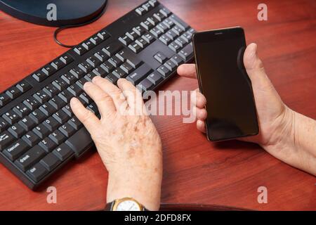 Mani di una donna anziana che scrive su un computer tastiera e contiene uno smartphone Foto Stock