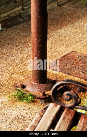 Dettaglio astratto di panca, lampada-palo e tombino, tutti arrugginiti, sul marciapiede - pavimentazione. Foto Stock