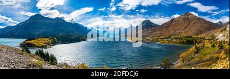 Upper Waterton Lake Lakeshore in autunno fogliame stagione soleggiata giorno mattina. Cielo blu, nuvole bianche sulle montagne sullo sfondo. Punti di riferimento Foto Stock