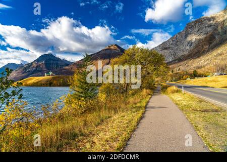 Middle Waterton Lake Lakeshore in autunno fogliame stagione soleggiata giorno mattina. Cielo blu, nuvole bianche sulle montagne sullo sfondo. Punti di riferimento Waterton Foto Stock