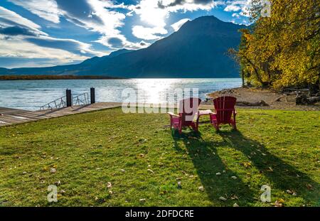 La sedia rossa guarda sopra il lancio della barca del lago di Waterton centrale nella mattina di stagione del fogliame autunnale. Luce del sole che passa il cielo blu e le nuvole su Mountains.Waterton Lake Foto Stock