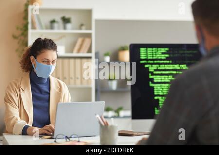 Sviluppatori in maschere che lavorano al tavolo sui computer con codici in ufficio Foto Stock