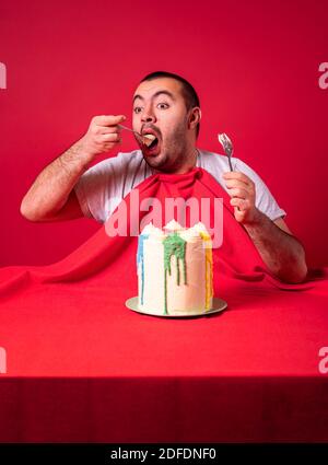 Giovane uomo che mangia tutto da solo enorme torta di compleanno. L'uomo affamato mangia la torta con entrambe le mani al tavolo su uno sfondo rosso. Foto Stock