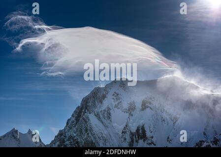 Forte vento soffia la neve sopra una cima di montagna creando una strana nuvola che guarda iridescente alla luce del sole contro un cielo blu profondo Foto Stock