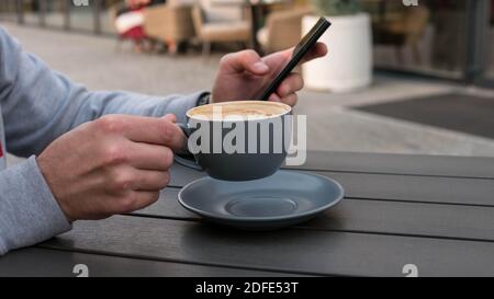 Bevande cappuccino e tipi al telefono, un giovane uomo gode di una tazza di caffè in un bar sulla strada, tenendo uno smartphone in mano. Foto Stock