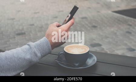Un giovane beve un cappuccino con uno smartphone, un uomo tiene un telefono in mano e beve caffè Foto Stock