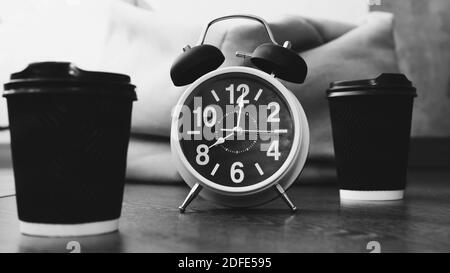 Orologio retrò con sveglia e cappuccino in tazze di plastica, orologio mani alle 8:00, caffè aromatico in tazze, immagine in bianco e nero, primo piano. Foto Stock