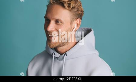 Bell'uomo elegante con la bearded in felpa con cappuccio che guarda felice parlare sopra auricolari wireless con amici su sfondo colorato Foto Stock