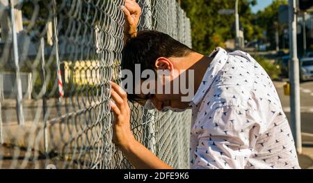 Uomo angosciato che piange e spinge la testa contro la recinzione. Vista profilo. Foto Stock