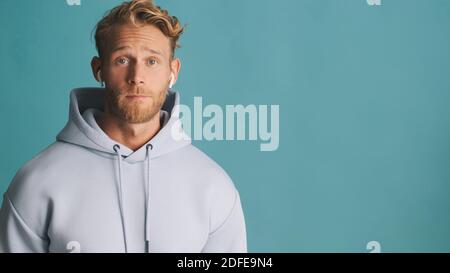 Elegante e attraente uomo in felpa con cappuccio indossa auricolari wireless in piedi vicino luogo per pubblicità o testo promozionale isolato su blu sfondo Foto Stock