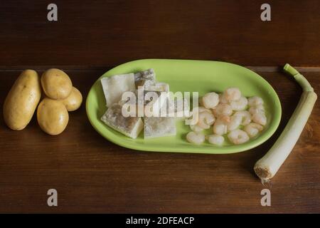 Un tavolo di legno con patate, porri e un piatto verde con filetti di merluzzo e gamberi. Ingredienti per cucinare pesce e verdure. Foto Stock