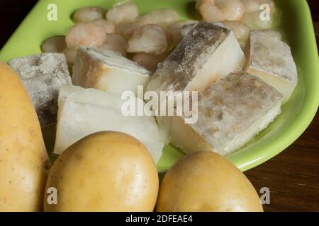 Alcuni filetti di merluzzo crudo con patate intere e gamberi sullo sfondo. Ricette di baccalà sane e cotte per la cottura a vapore con ingredienti naturali. Foto Stock