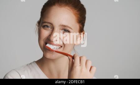 Ragazza sorridente attraente che guarda felicemente in macchina fotografica spazzolando i denti sopra sfondo bianco Foto Stock