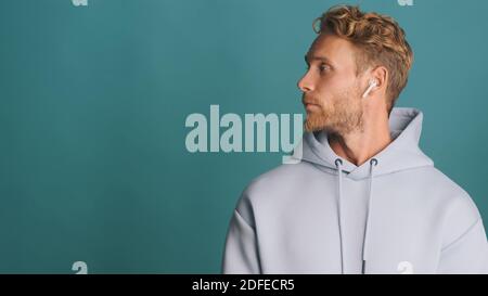 Vista laterale dell'elegante uomo con portacolori in felpa con cappuccio senza fili auricolari in piedi vicino al posto per pubblicità o testo promozionale sfondo blu Foto Stock