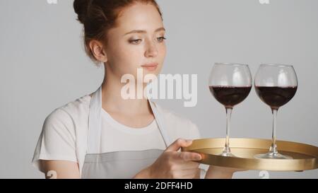 Cameriera giovane in grembiule che tiene intentemente il vassoio con il vino sopra sfondo bianco Foto Stock