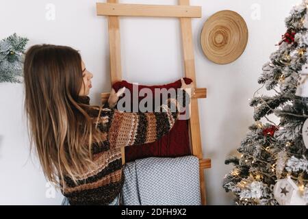 Giovane donna decora casa per Natale, appende plaid su scala di legno vicino albero di Natale. Foto di alta qualità Foto Stock