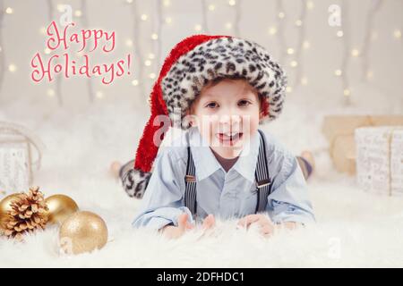 Buone vacanze. Cartolina di Natale con testo. Carino adorabile ragazzo caucasico in cappello di Santa sdraiato su un soffice tappeto bianco a terra. Tradizionale Natale New Y Foto Stock