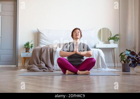 Una donna anziana medita a casa nella posizione di loto. Lezioni di yoga per prolungare i giovani. Concetto di stile di vita sano. Foto Stock