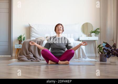 Donna anziana medita a casa nella posizione di lotus. Lezioni di yoga per prolungare i giovani. Stile di vita sano e concetto anti-età. Foto Stock