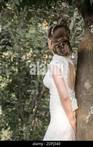 Una sposa in un abito bianco si erge su un albero nel Parco, vista posteriore Foto Stock