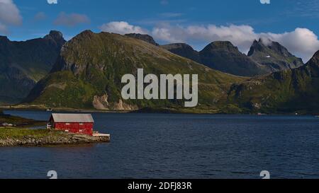 Idilliaca vista di piccolo capannone con la facciata dipinta di rosso situato sulla riva del tranquillo fiordo Selfjorden con le splendide e aspre montagne di Flakstadøya. Foto Stock