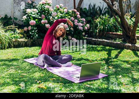 Sport online fitness yoga allenamento di formazione, donna anziana nel giardino della sua casa facendo esercizi su pilates tappetino di fronte computer portatile a casa. Foto Stock