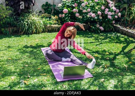 Sport online fitness yoga allenamento di formazione, donna anziana nel giardino della sua casa facendo esercizi su pilates tappetino di fronte computer portatile a casa. Foto Stock