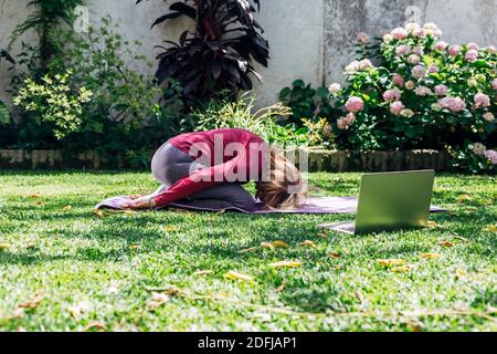 Sport online fitness yoga allenamento di formazione, donna anziana nel giardino della sua casa facendo esercizi su pilates tappetino di fronte computer portatile a casa. Foto Stock