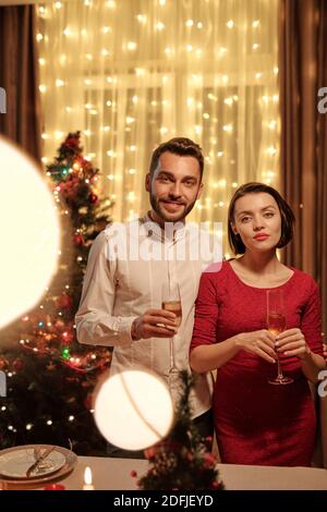 Giovane bella donna in abito rosso e suo marito tostare con flauti di champagne mentre si sta in piedi al tavolo fesive contro l'albero di natale Foto Stock